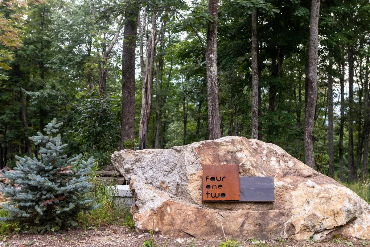 A large rock engraved with a sign welcomes guests at the lodge entrance, surrounded by lush greenery and evergreens. The photo captures the first impression upon arrival and hints at easy access to nearby trails and natural beauty.