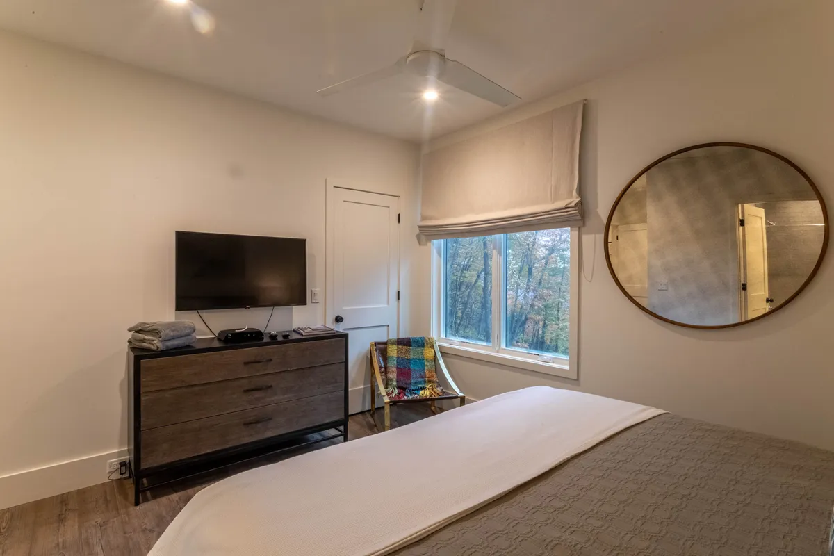 A comfortable and serene guest room featuring a bed, window with soft natural light, a dresser with TV, round mirror, and a colorful chair. The room exudes a feeling of privacy and relaxation, suited for guests like grandparents or friends.