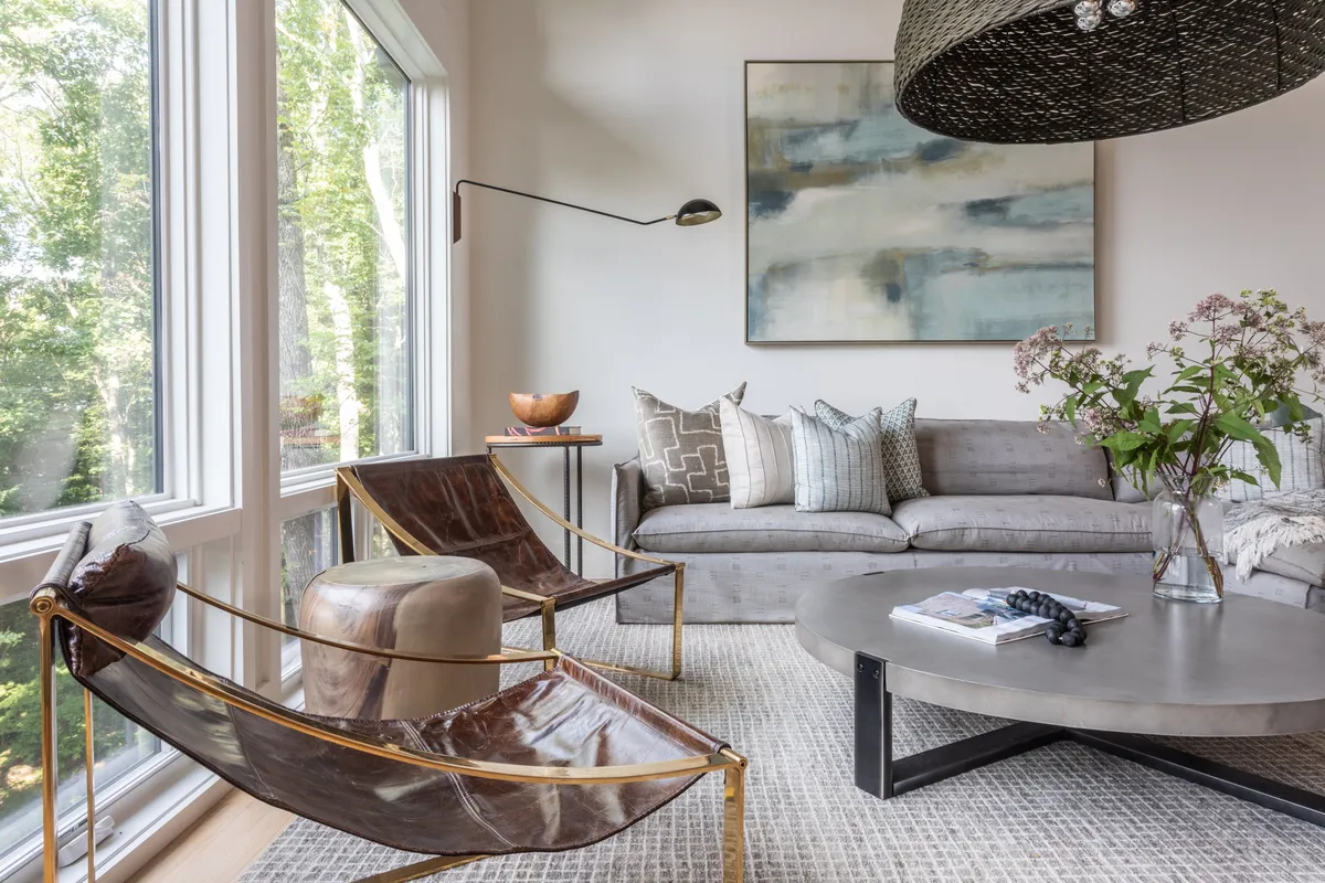 This image captures a great room with large windows letting in ample natural light, stylish seating, modern artwork, and a glass vase with flowers. The scene is welcoming and tranquil, with cozy seating arranged to encourage conversation and relaxation while taking in scenic views.