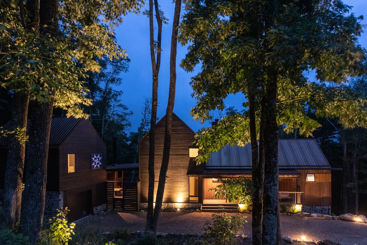 The exterior shot shows the inviting wooden facade of the lodge nestled among mature trees, with soft landscape lighting illuminating the entrance. The tranquil setting radiates a sense of seclusion and connection to nature, ideal for unwinding or transitioning between activities.