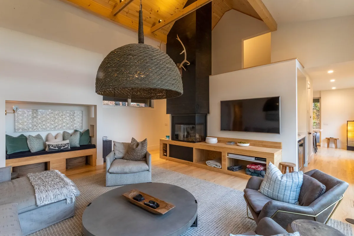 A cozy, welcoming seating area is centered around a modern wood-burning fireplace, complemented by a large woven pendant light and comfortable couches. Wooden accents and soft cushions evoke warmth and togetherness, making this space perfect for gatherings.