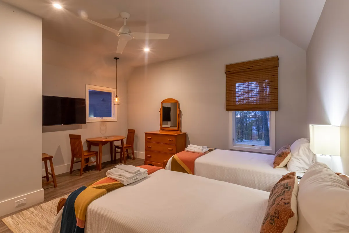 A spacious, multipurpose room with two beds, a dining table set for games or gatherings, a dressing mirror, and a window view. The room suggests flexible uses—from family board game tournaments to casual relaxation.