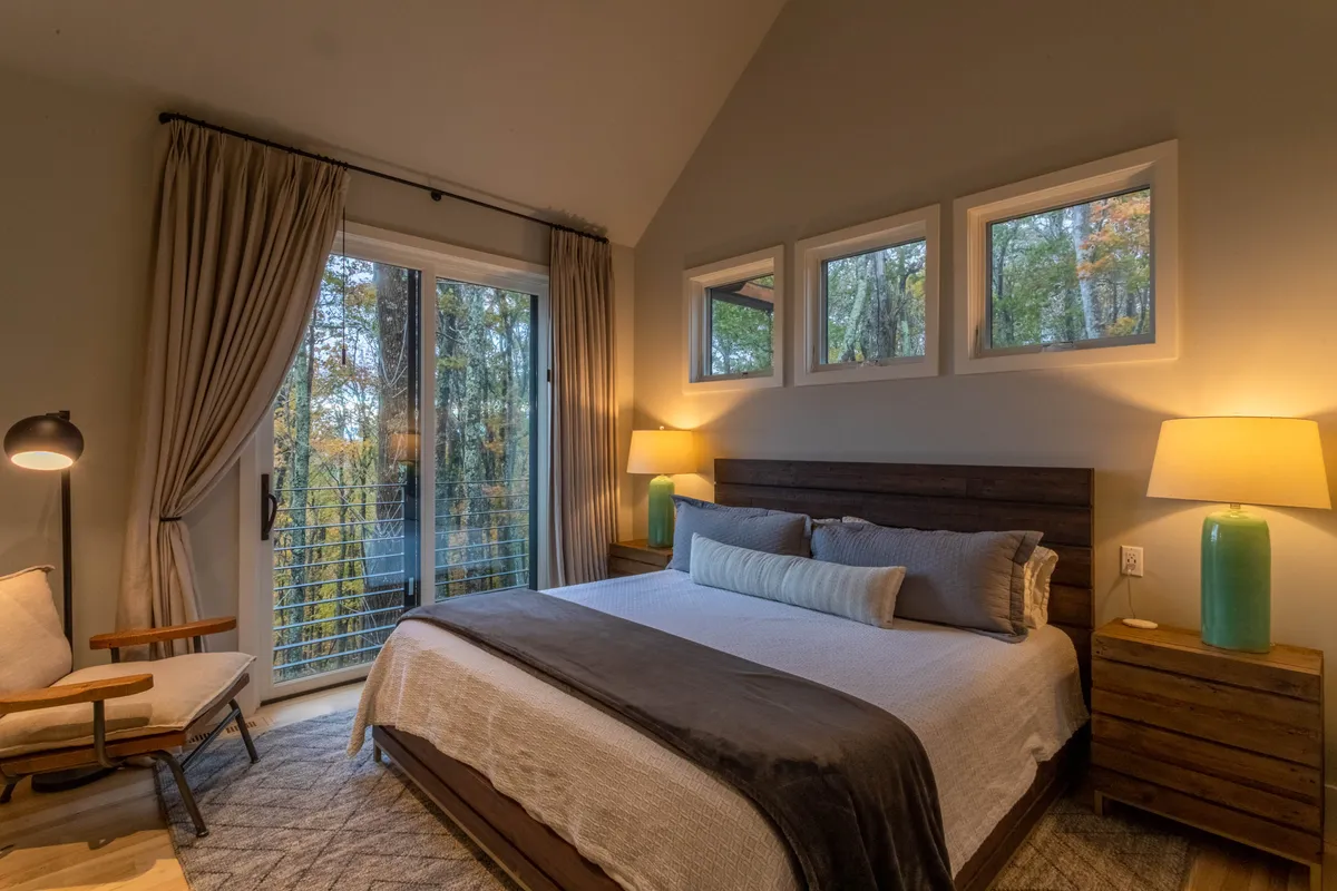 A serene and inviting master suite featuring a bed with textured bedding, large windows offering a nature view, and a cozy nightstand with a lamp. The overall design highlights high-end finishes and a tranquil atmosphere.