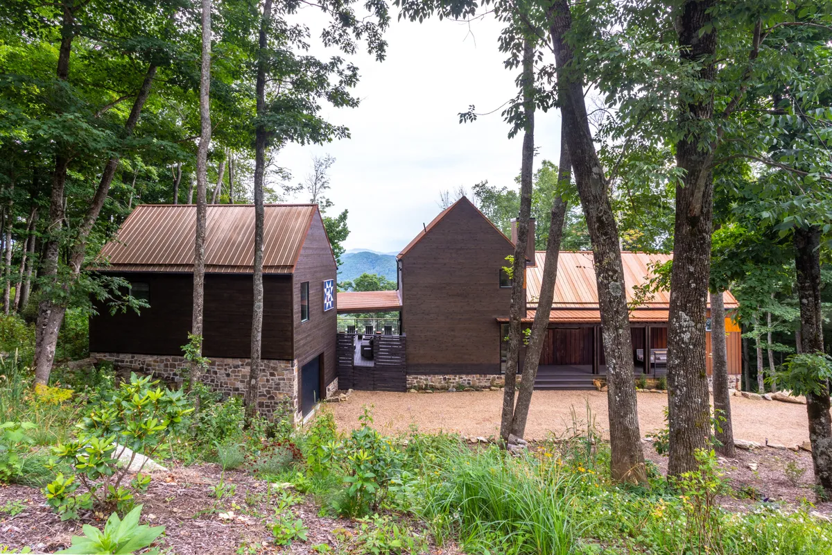 This image captures two distinct, architecturally striking buildings set against a serene mountain backdrop, surrounded by natural greenery. The separation of the structures hints at the presence of detached guest quarters, while the open setting emphasizes privacy and exclusivity. The scene is bathed in sunset light, reinforcing the mountain retreat vibe.