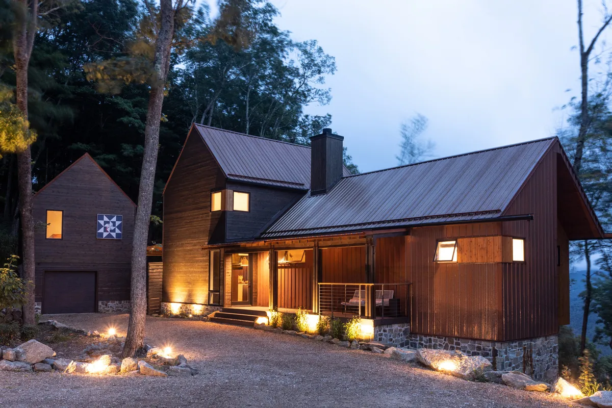 The hero image showcases the striking modern mountain architecture of Four One Two Lodge, featuring natural stone and wood elements warmly illuminated at dusk. A landscaped pathway leads to the inviting porch, where cozy lighting hints at evenings spent by the outdoor fireplace, all set against the tranquil backdrop of the Blue Ridge Mountains.