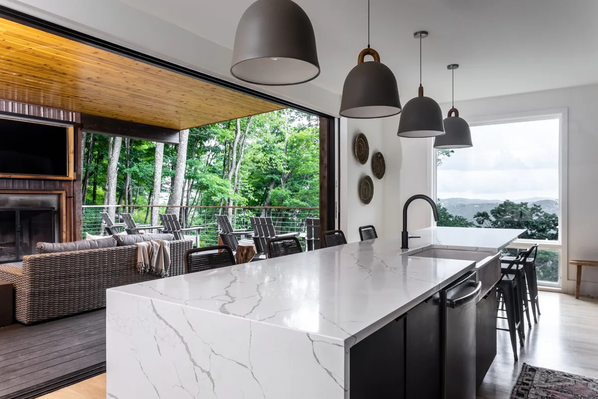 A panoramic view of a contemporary kitchen with a large island, hanging pendant lights, and a stunning natural backdrop seen through wide windows. The space is designed for both cooking and entertaining, blending seamlessly with the living area for a modern open-concept feel.
