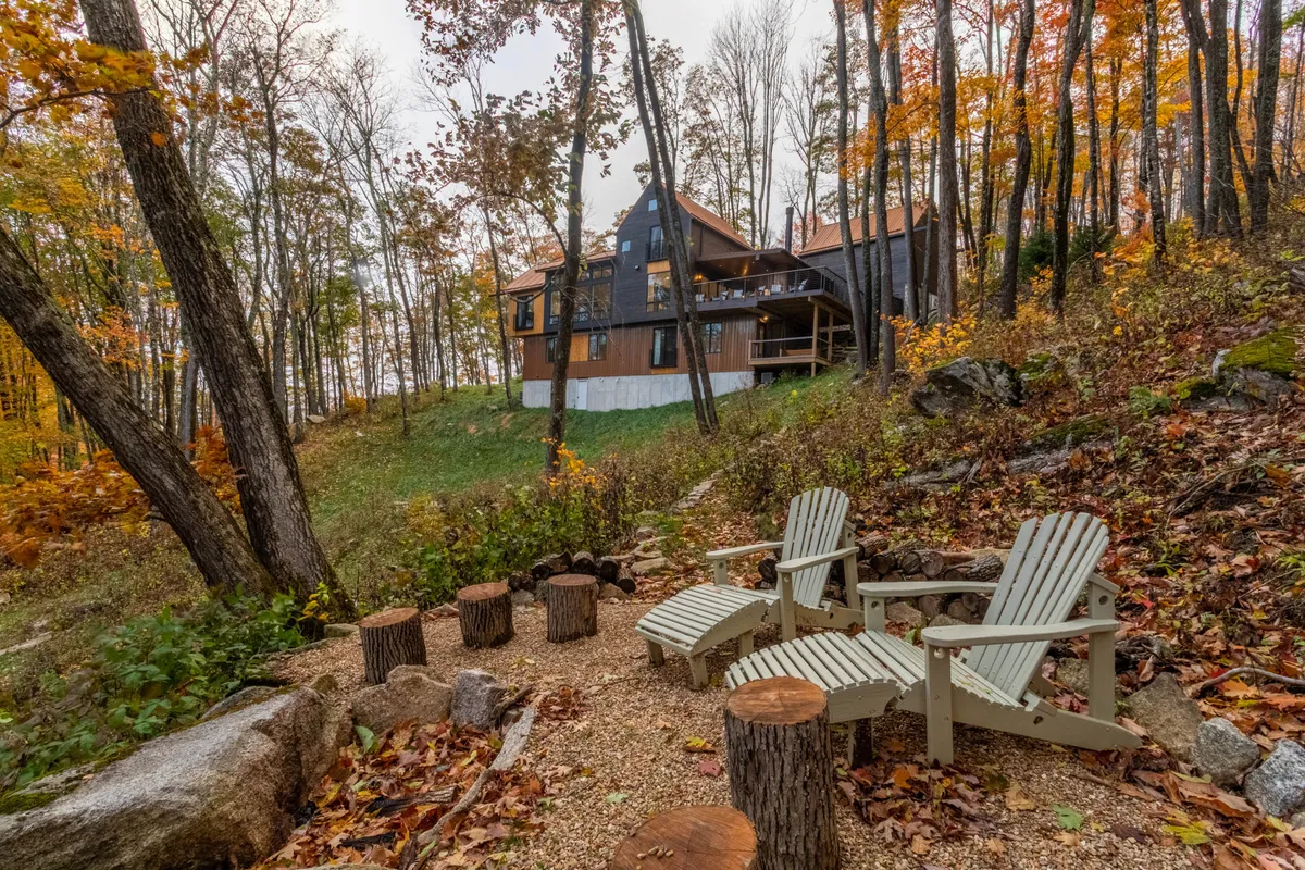 A cozy outdoor lounge scene with wooden chairs arranged around logs, set before the house and surrounded by vibrant fall foliage. The setting is ideal for sunset views, relaxing with a drink, or enjoying an evening campfire, with the house providing a luxurious backdrop.