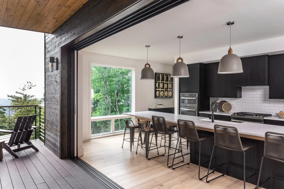 An airy kitchen and great room with a large open window and an island counter, allowing light and views from outside to flow into the space. The wooden deck just outside further emphasizes the connection to nature, reinforcing the sense of openness and inviting atmosphere described in the section.