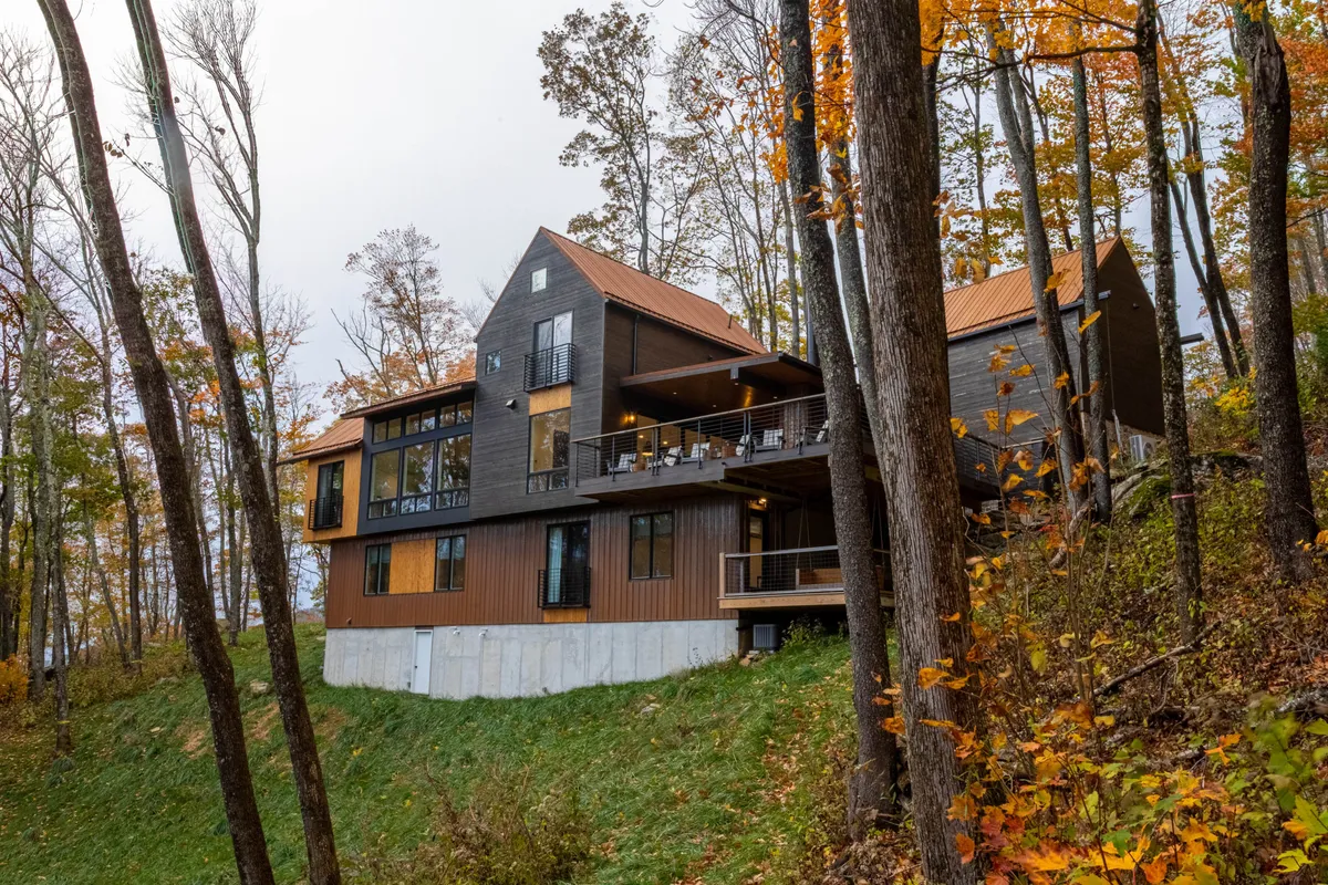 The exterior of a modern lodge featuring spacious decks, wood and stone architecture, large windows, and inviting outdoor gathering areas nestled among trees. The deck is set up for relaxation and family barbecues, linking indoor and outdoor living.